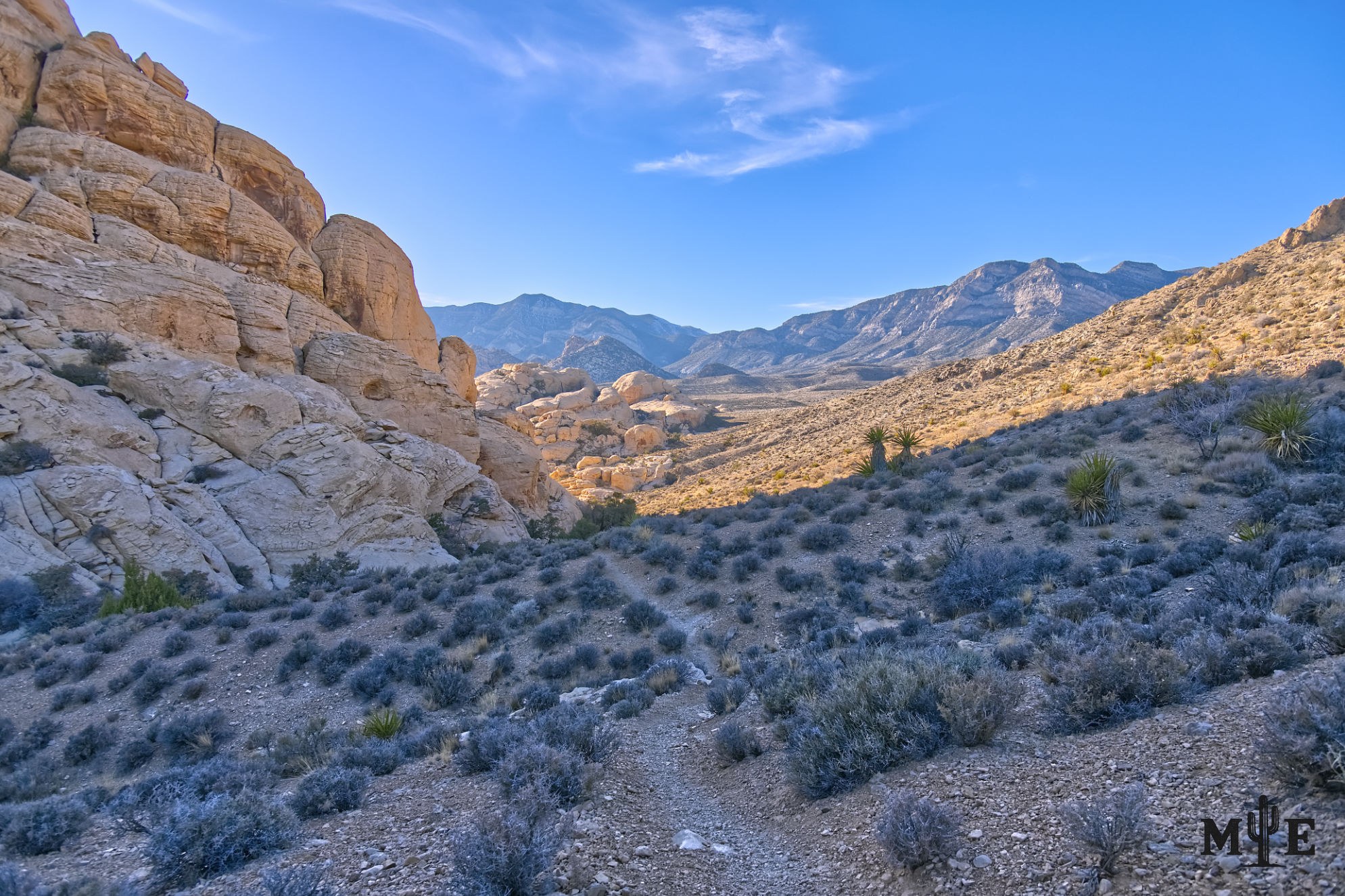 Hiking trail cutting through desert terrain in late afternoon under a blue sky with vast shadows cast from mountainsides.