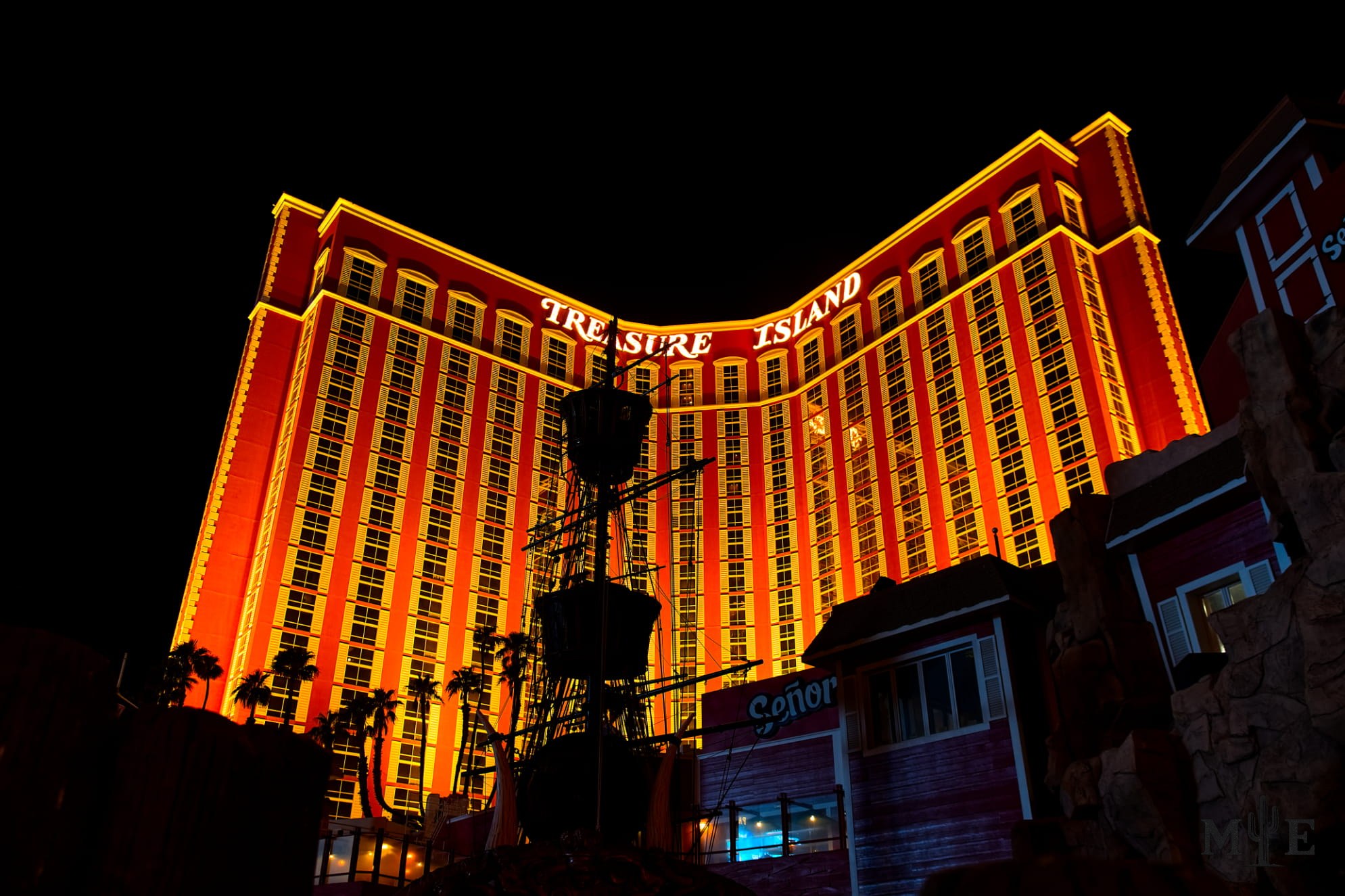 Stark contrast of the dark night sky and Treasure Island hotel and casino, illuminated in red orange with foreground shadows.