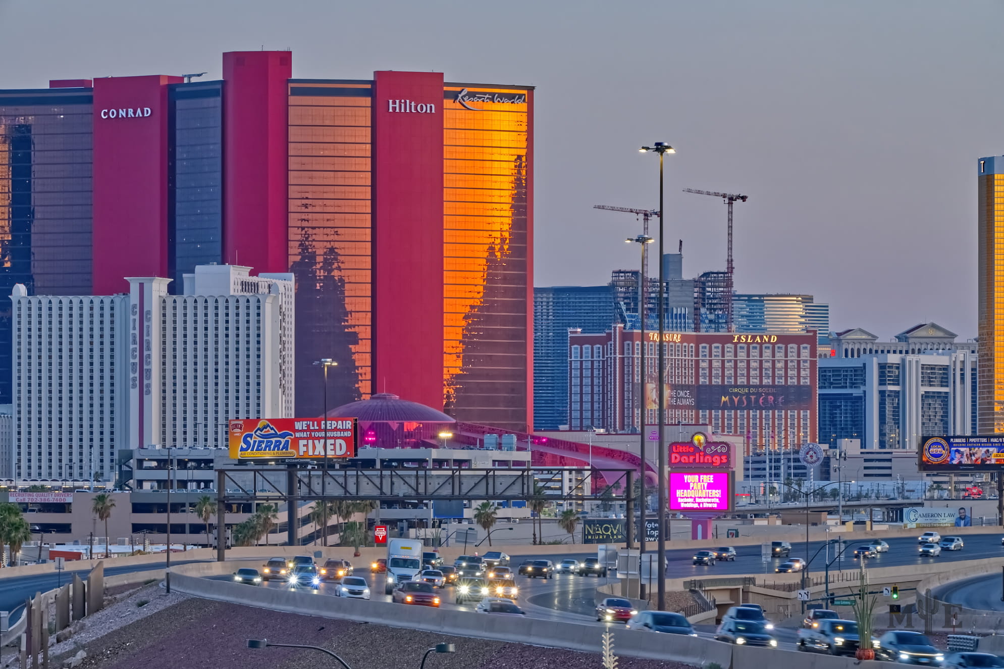 Hilton hotel glimmers at sunset adjacent to the freeway. Light grey sky and cars with headlights moving at dusk.