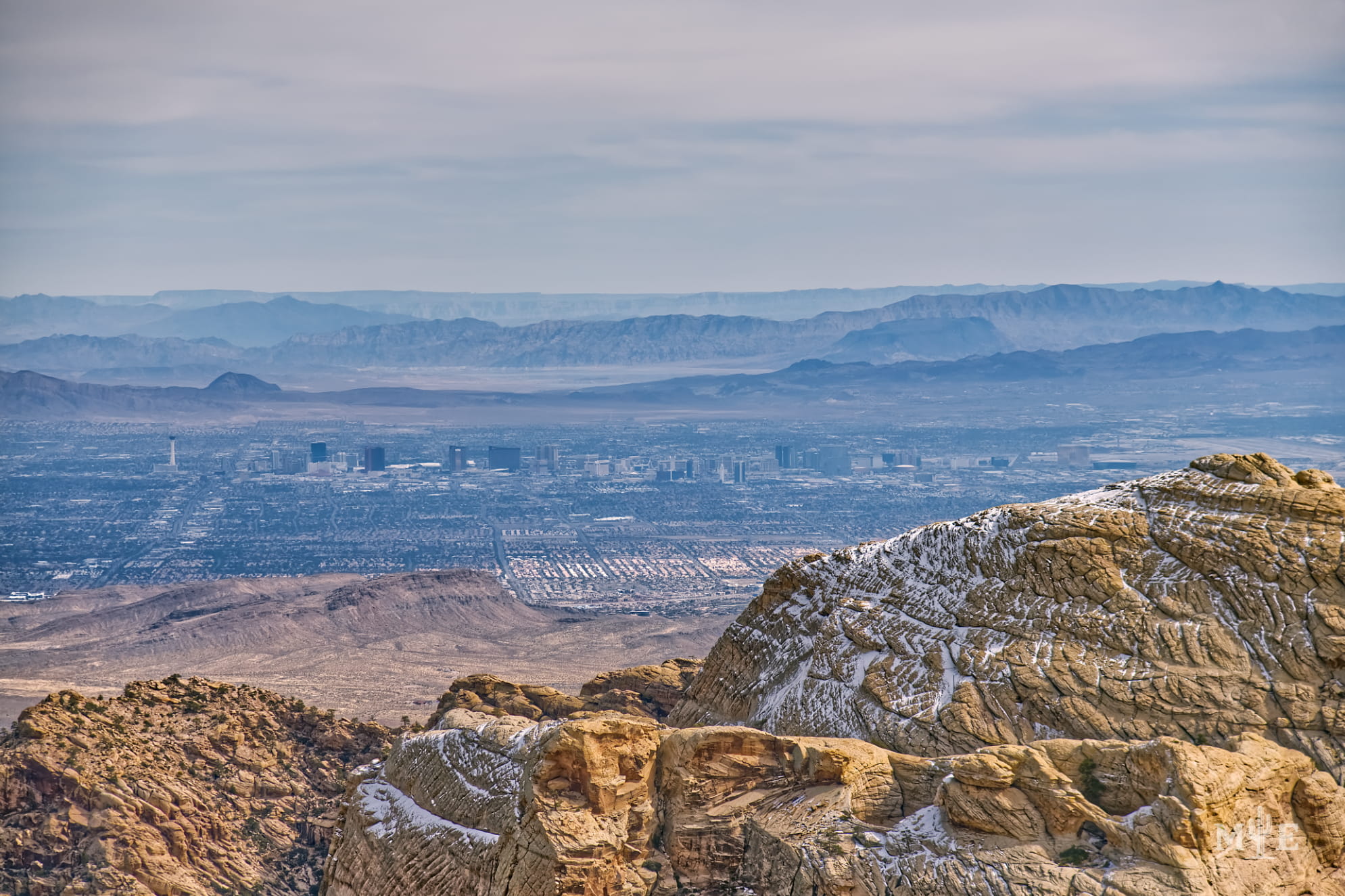 Lightly covered snow capped sandstone mountain in foreground with the city of Las Vegas, distant mountains and clouds.