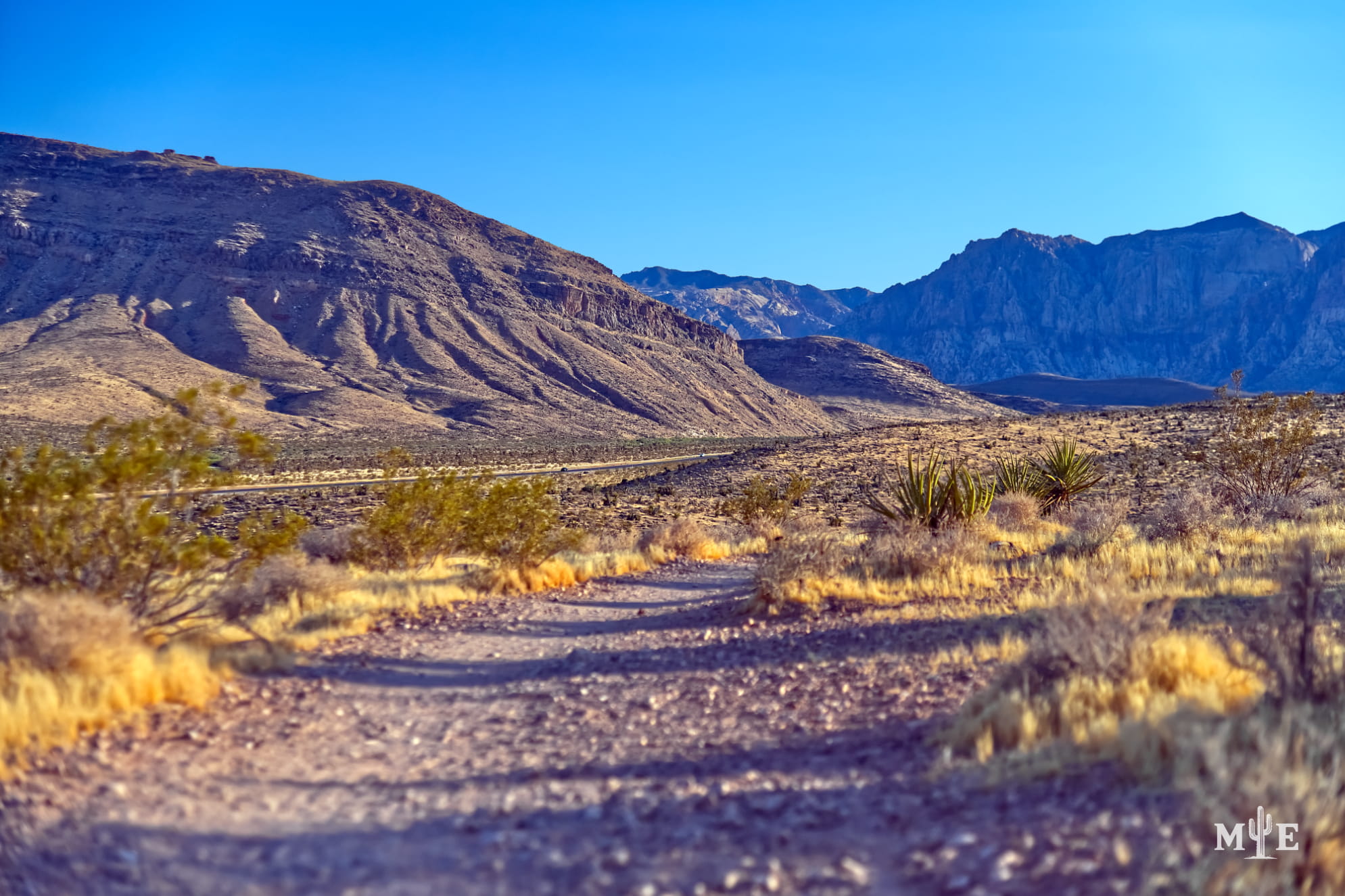 Blue sky over medium sized mountains in Southern Nevada, Mohave Desert terrain, and a trail front and center.