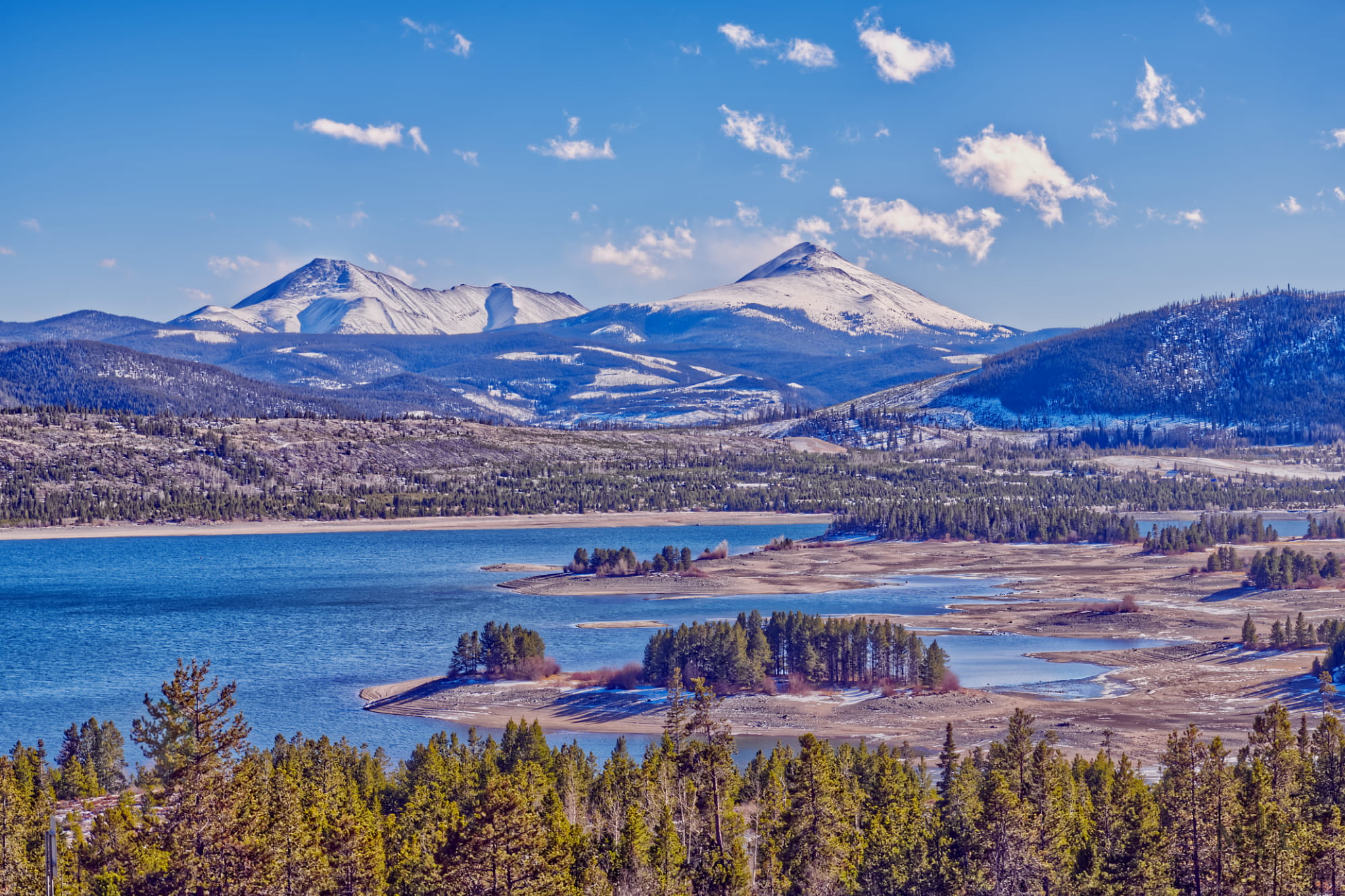 Gorgeous winter photograph of the Colorado Rockies, cold blue sky, and fresh water lake with lush green pine trees.