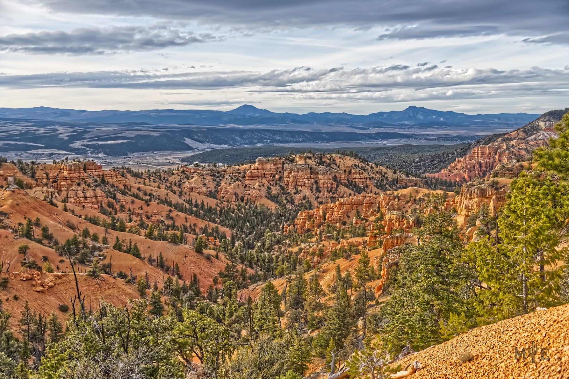 Scattered soft clouds over a rugged orange-gold canyon in Red Canyon Utah. Darker colored mountains in the distance.