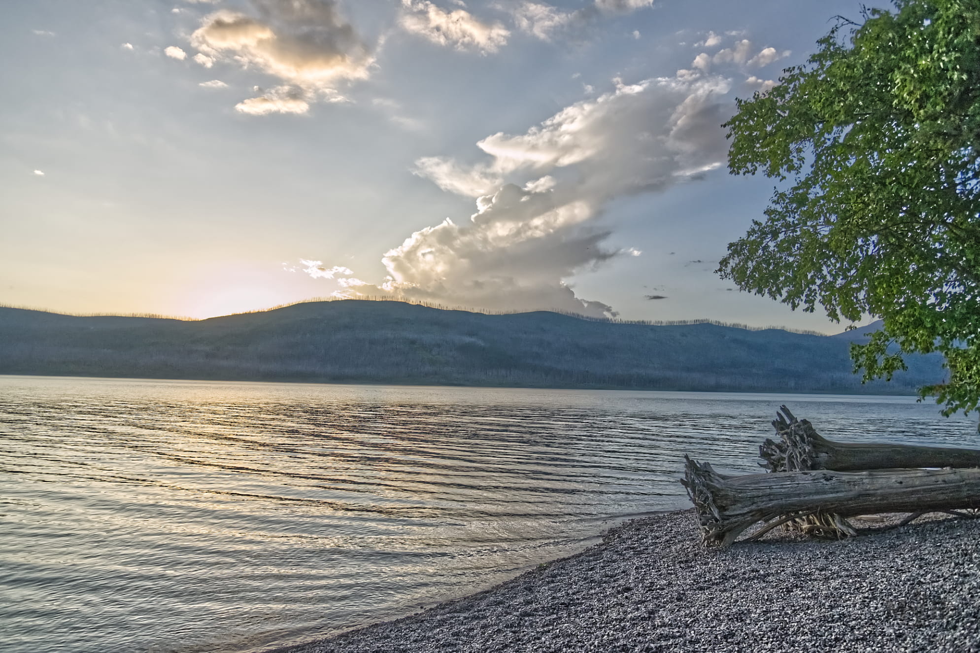 Gravel lake shore with calm and reflective water. Moutains delinate the clouds with subtle lake sunset.