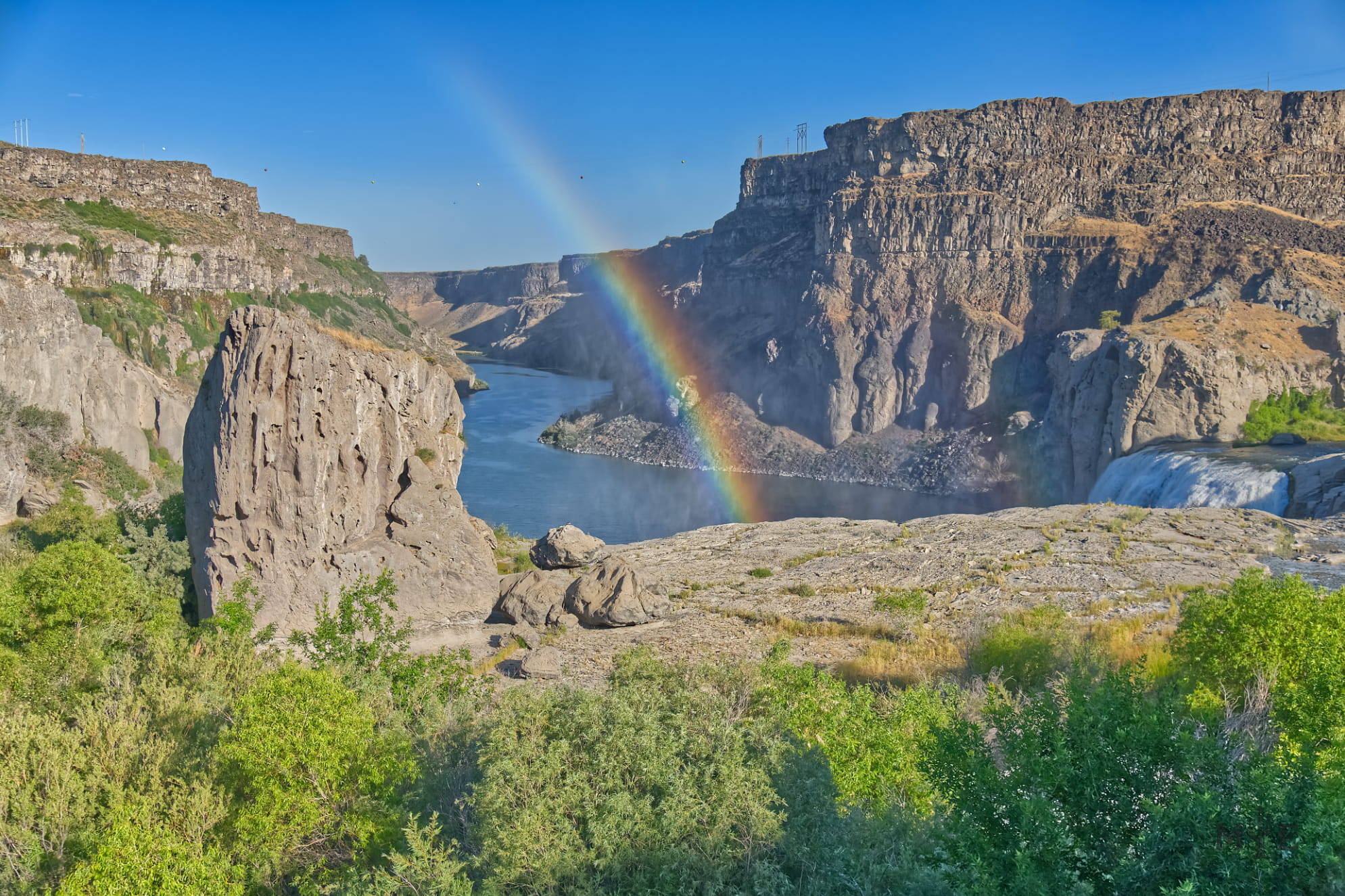 Rainbow diving into the river at Shoshone Falls in Idaho. Rugged mountains and dense green foliage surround.