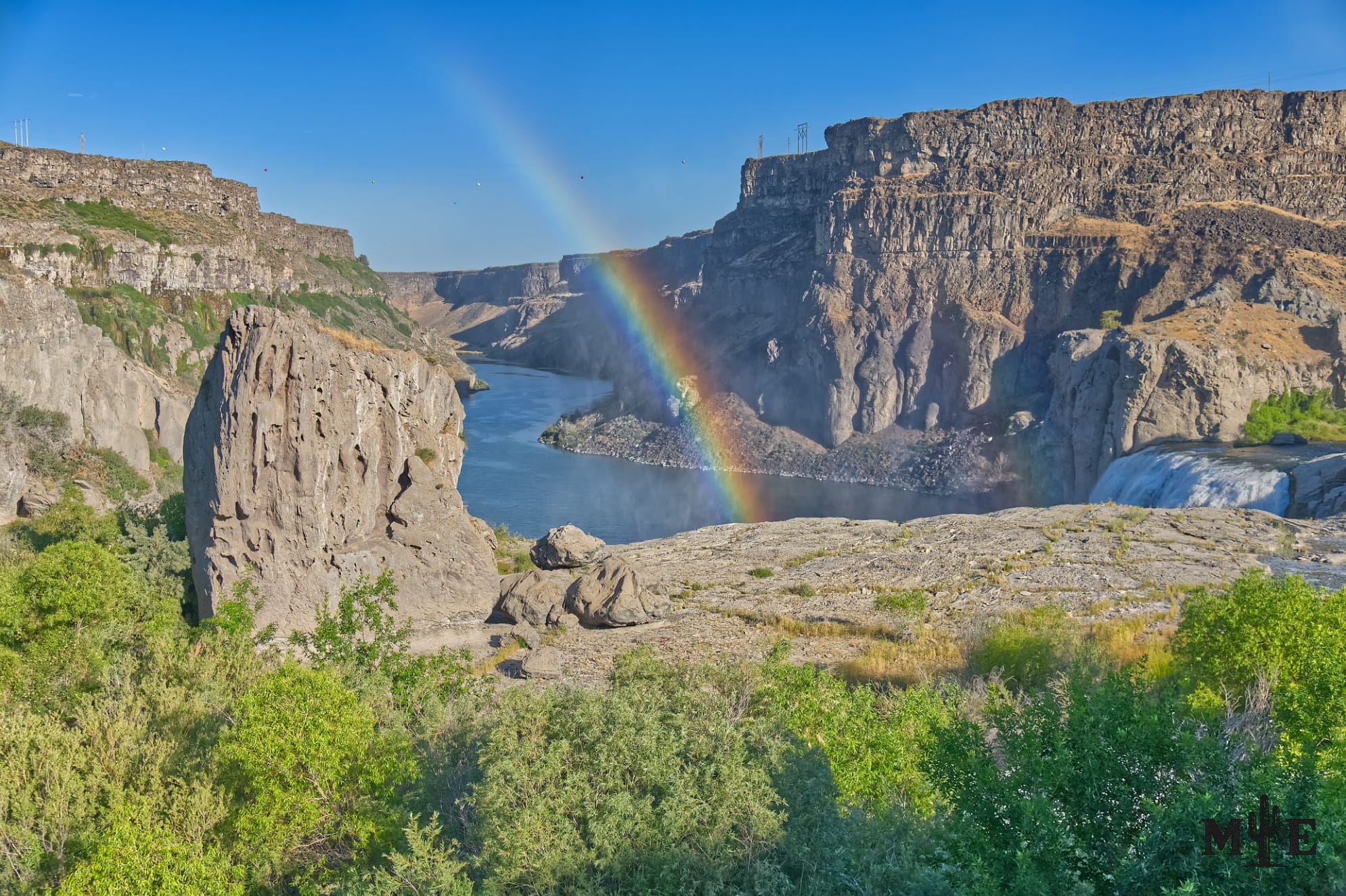 A rainbow descending toward the Snake River below Shoshone Falls, surrounded by rugged canyon walls and dense green vegetation.