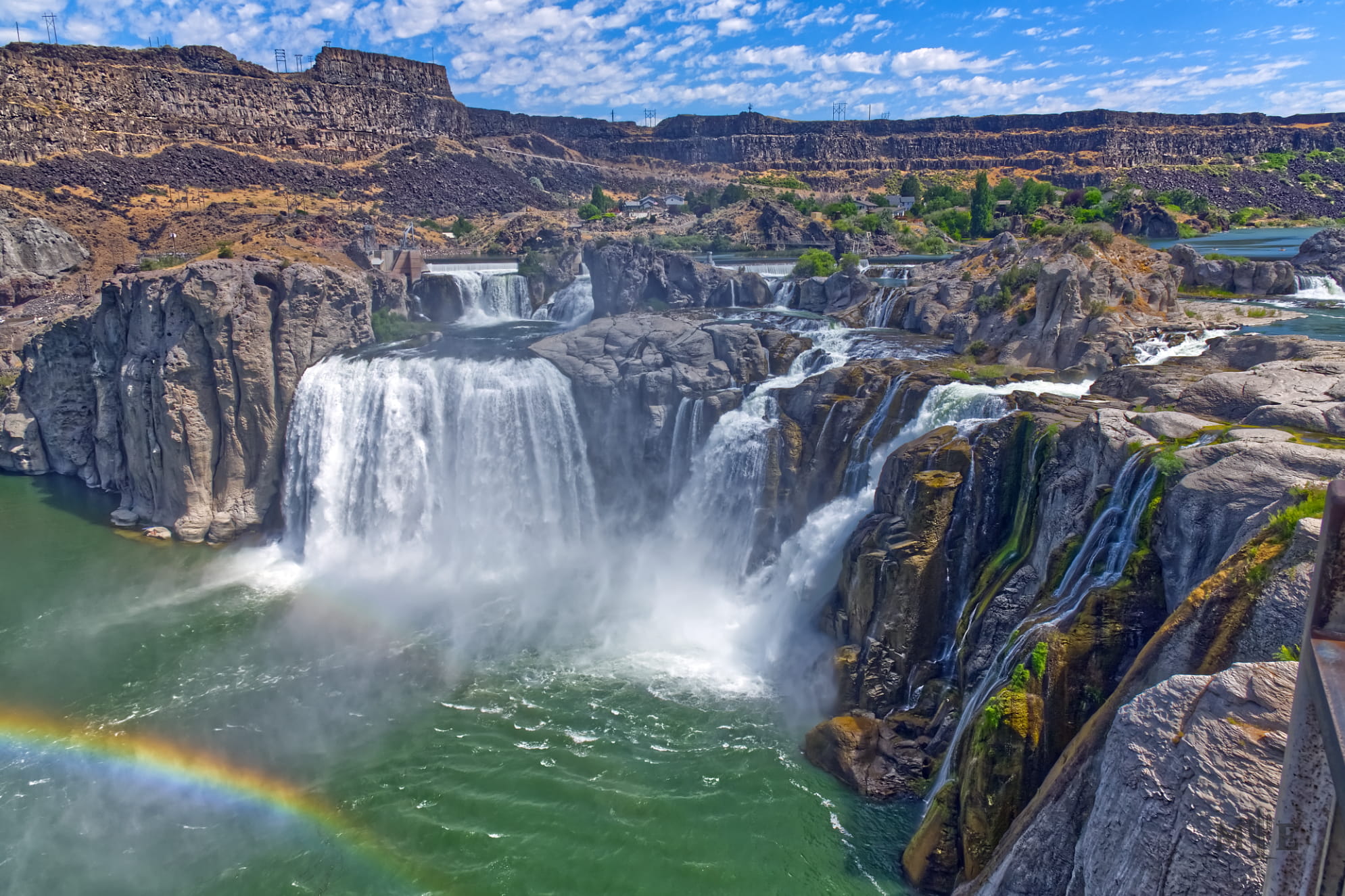 Blue sky over a series of powerful waterfalls at Shoshone Falls Idaho. Emerald green water and a rainbow at the bottom.