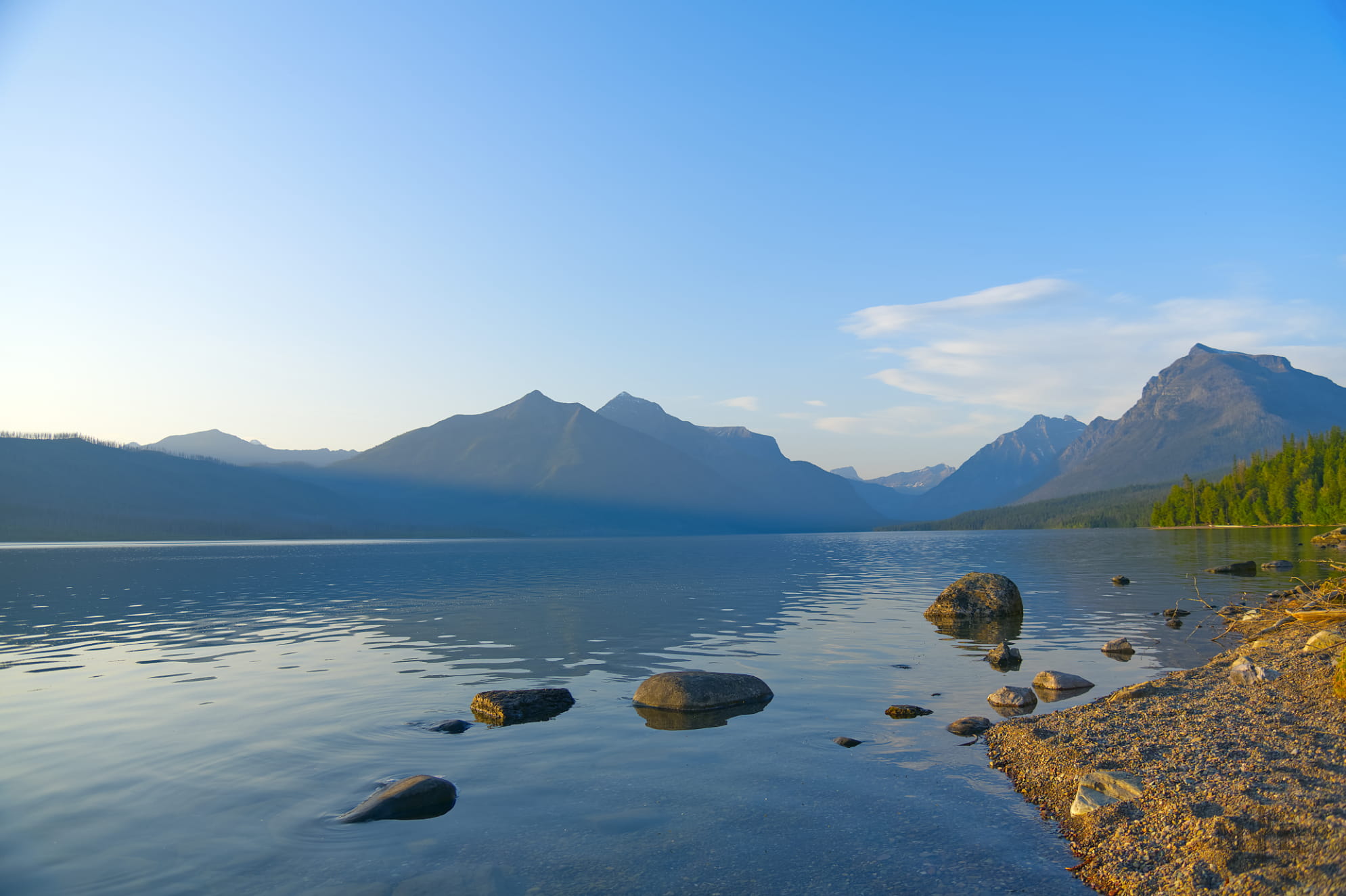 Calm and reflective lake at Glacier National Park with peaceful blue sky and mountains in the background.