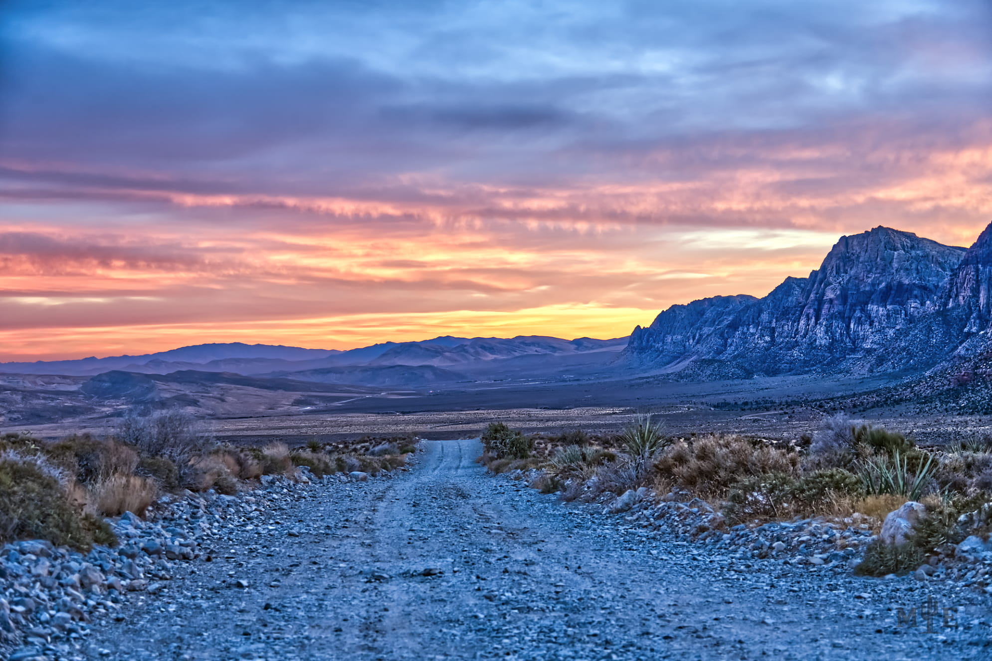 Gravel mountain road leading to a colorful desert sunset with colors ranging from yellow and pink to grey and blue.