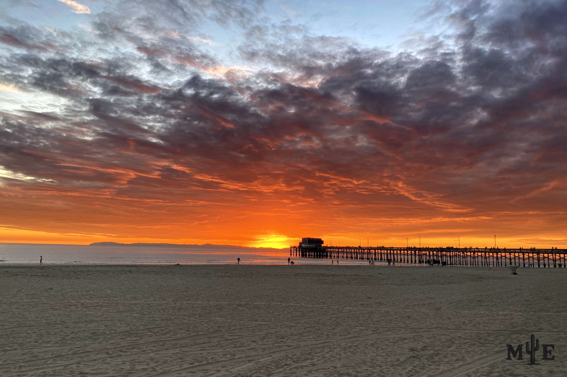 Newport Beach sunset with a few silhouetted figures on the sand beneath yellow, orange and red light fading into a blue-gray sky.