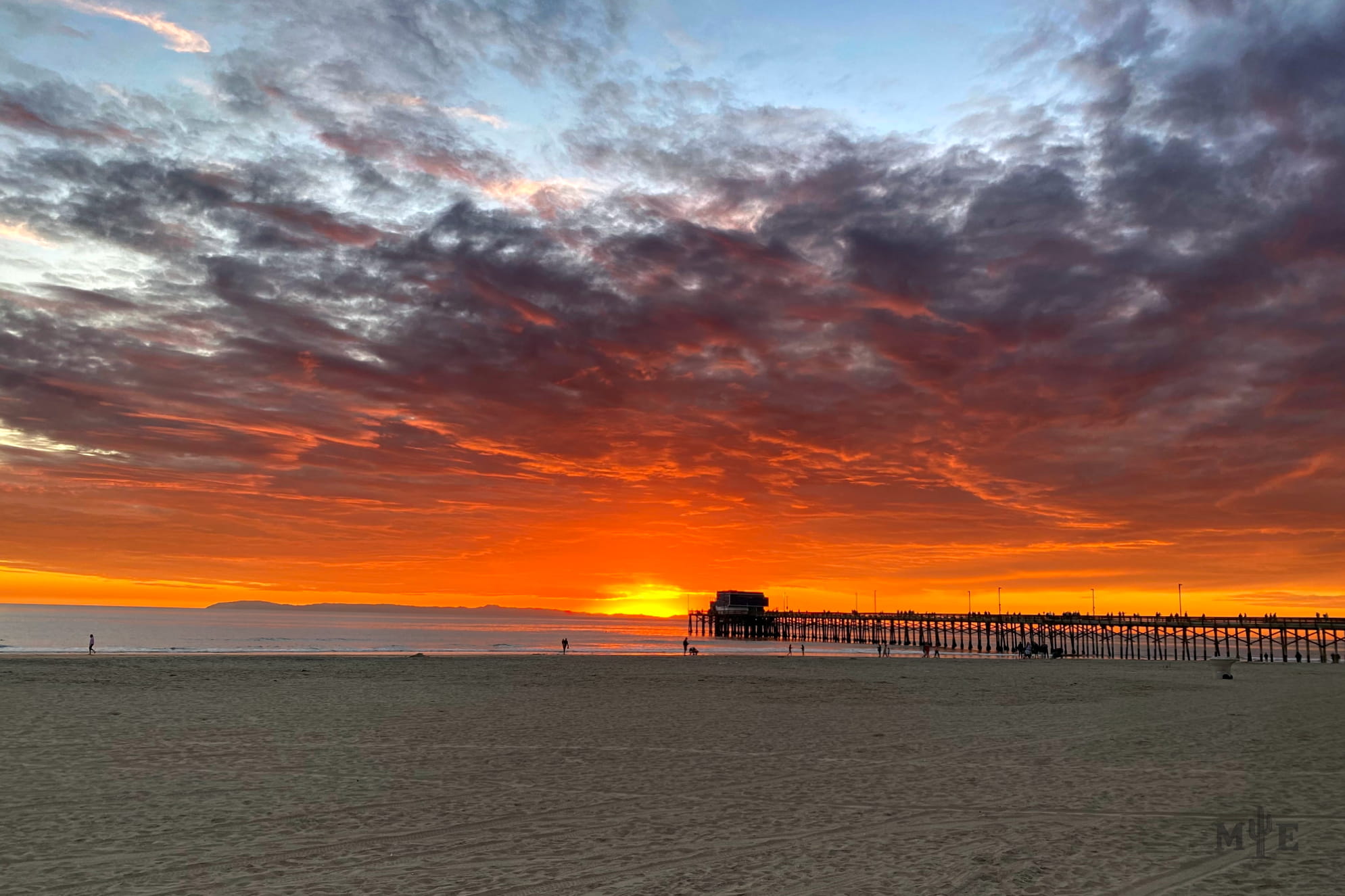 Newport Beach sunset with but a few silhouettes on the sandy beach. Yellow, orange, red, dark grey to light blue sky.
