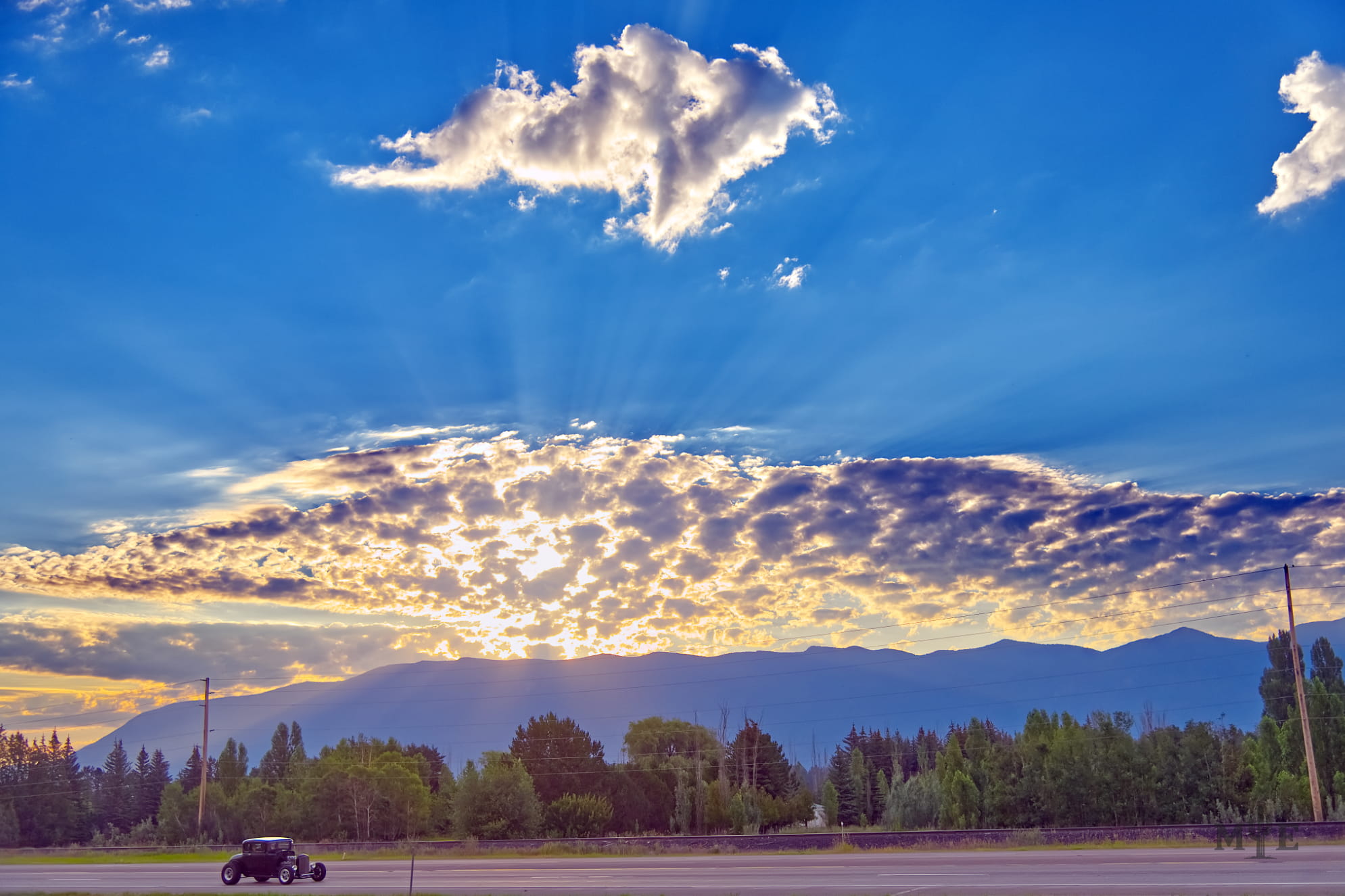 Sun rays bursting from clouds creating a wonderous sunset over a highway carrying a 1930s era Ford coupe in rural Montana.