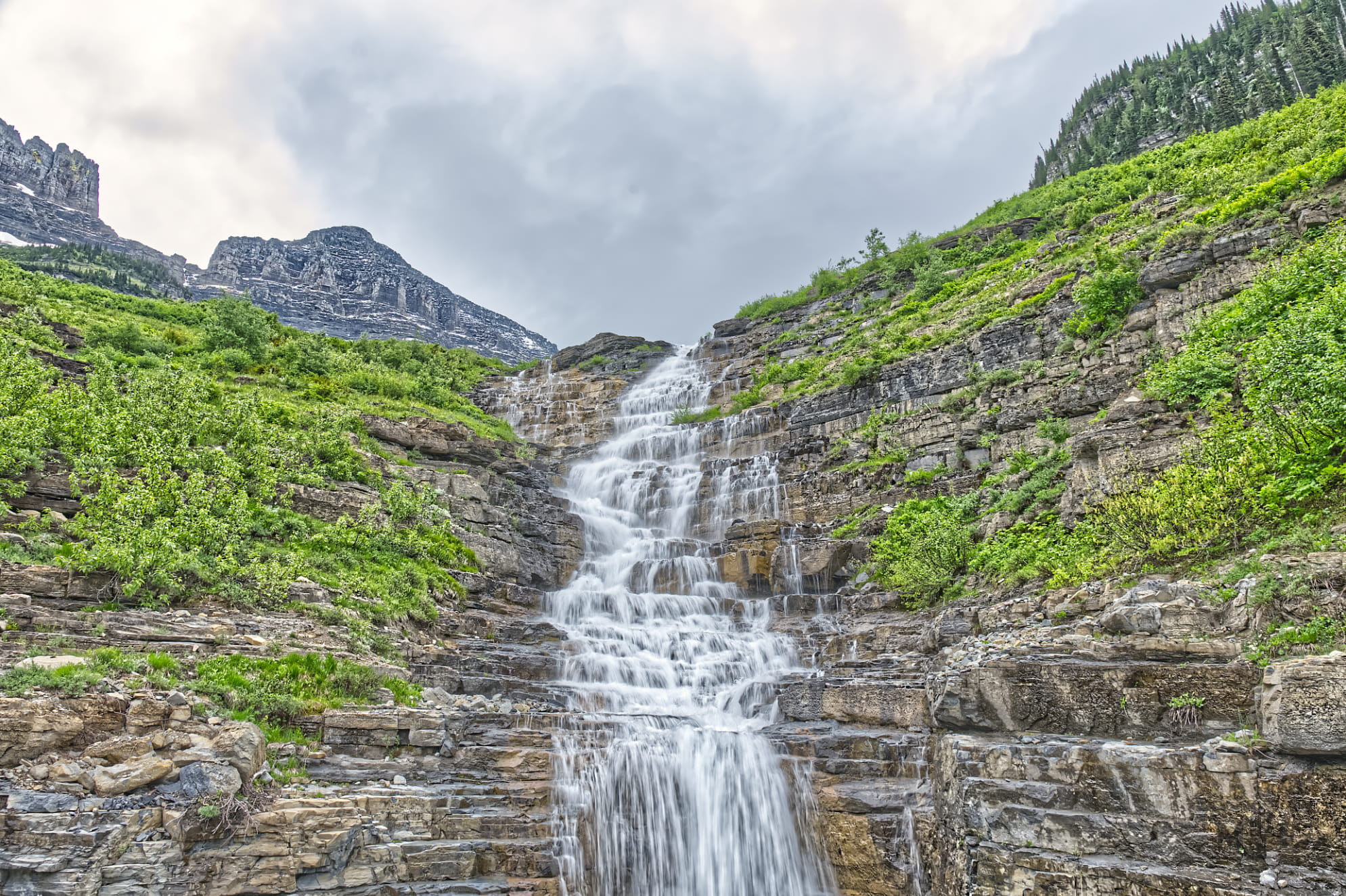 Long exposure waterfall in Glacier National Park moving over a stepped rock mountain landscape with overcast sky.