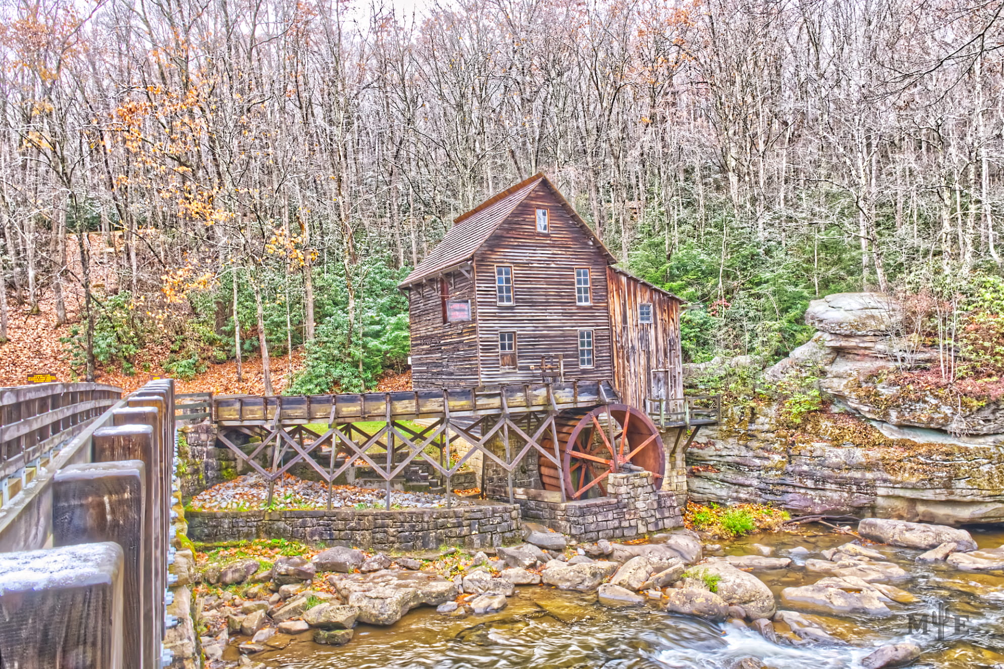 Historic mill on a beautiful creek with a bridge on the far left side. Dense foliage covers the background.