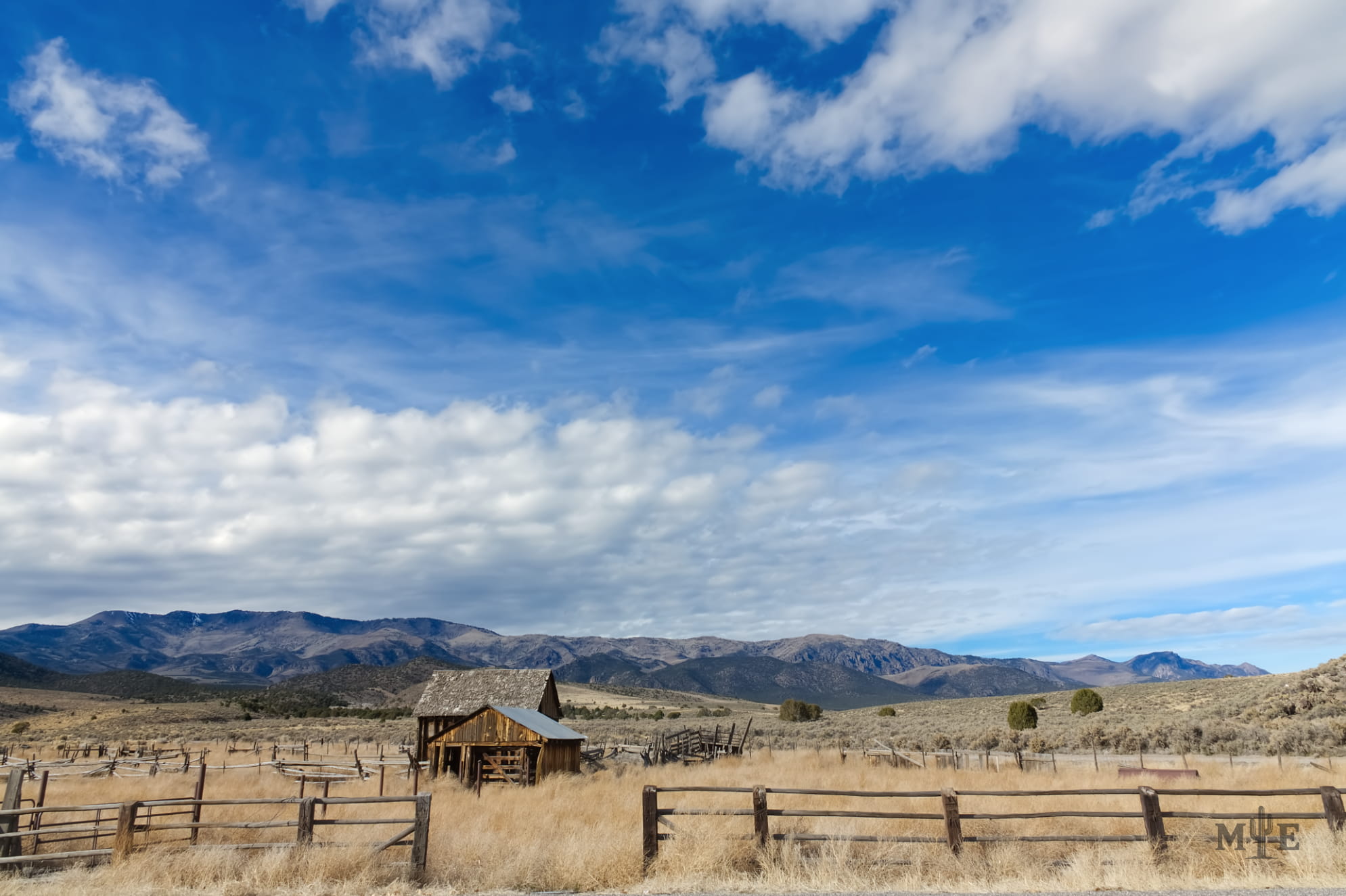 Blue sky with pastel clouds scattered about. Dried brush and old barn or farmhouse front and center. Wooden fence at bottom.