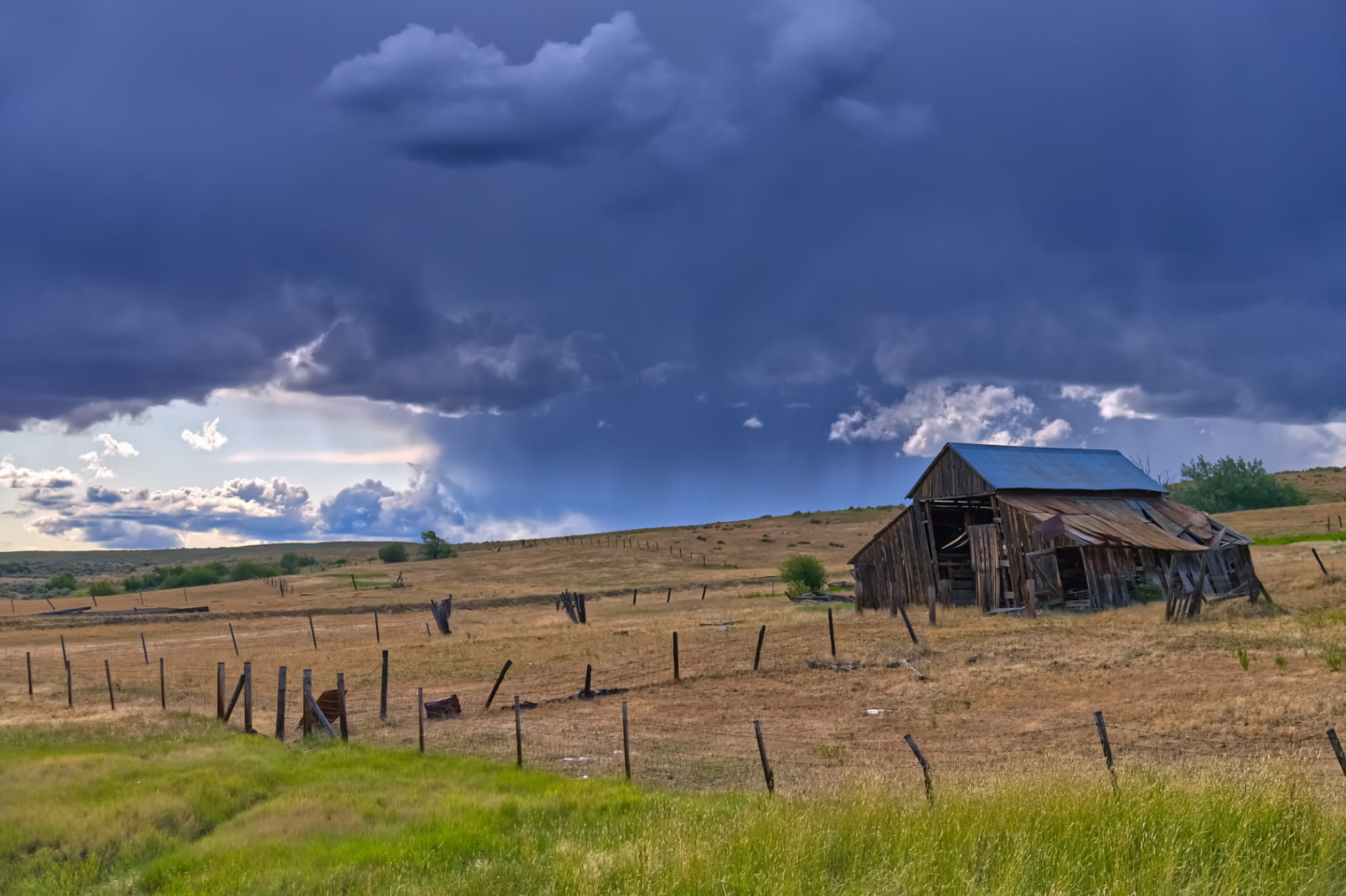 Ominous blue grey clouds covering an abandoned old barn/ farmhouse sitting atop of dried brush and green overgrown grass.