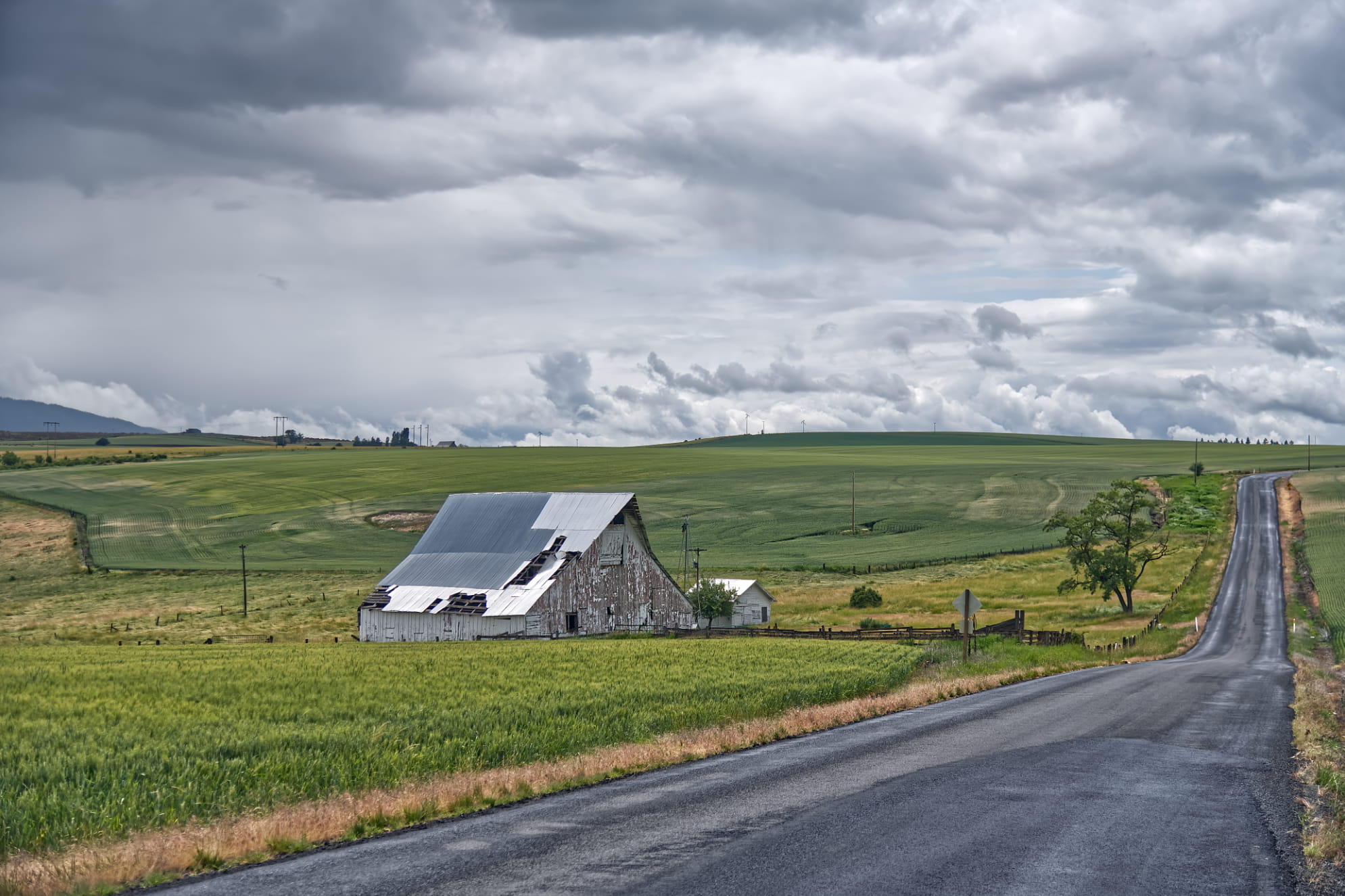 Long empty road in middle America with green farmland for as far as the eye can see with old barn rising up from the scene.