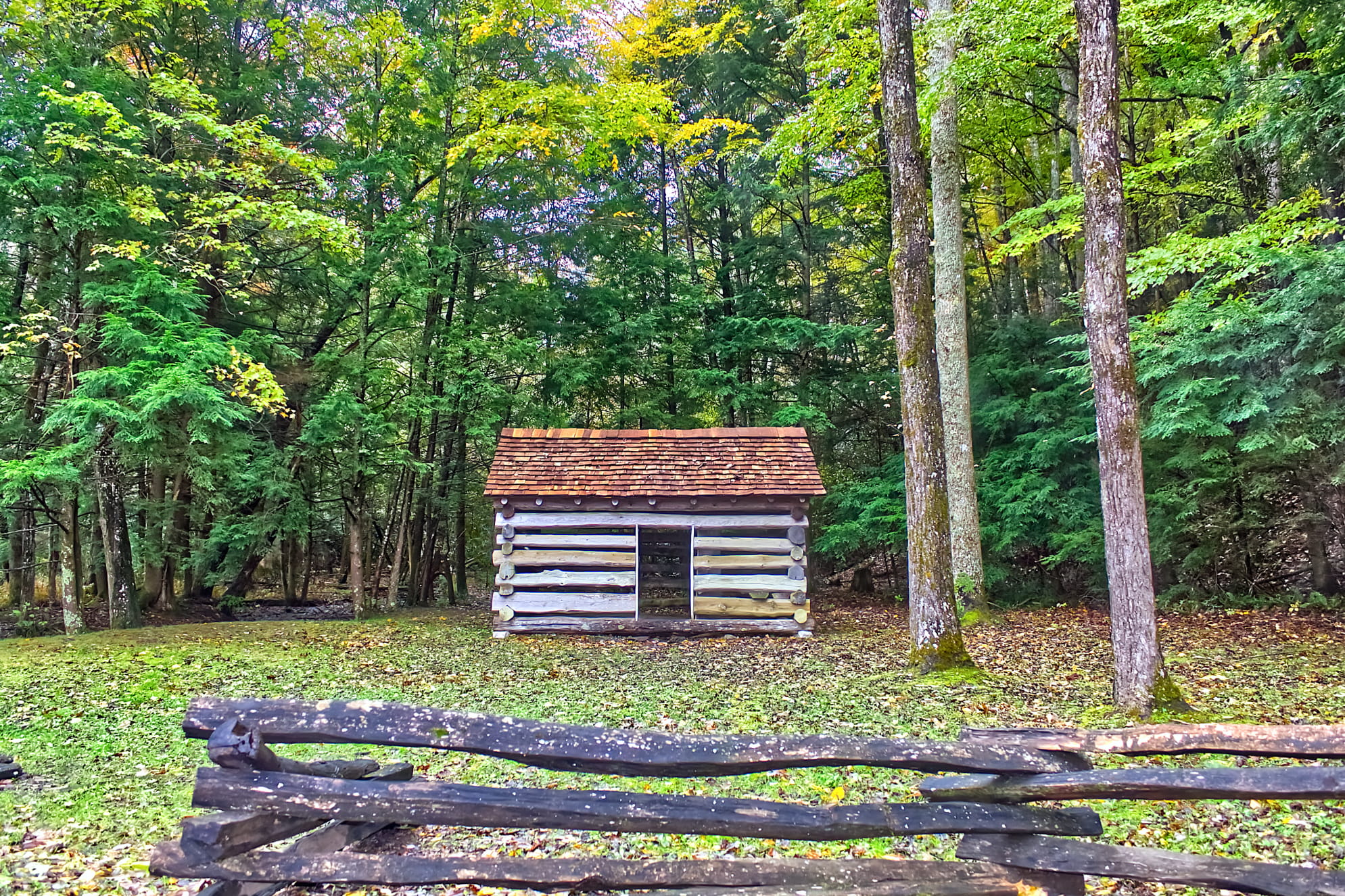 1800's era wooden fence with historic log cabin front and center. Surrounded by dense lush green foliage. Rural dwelling.
