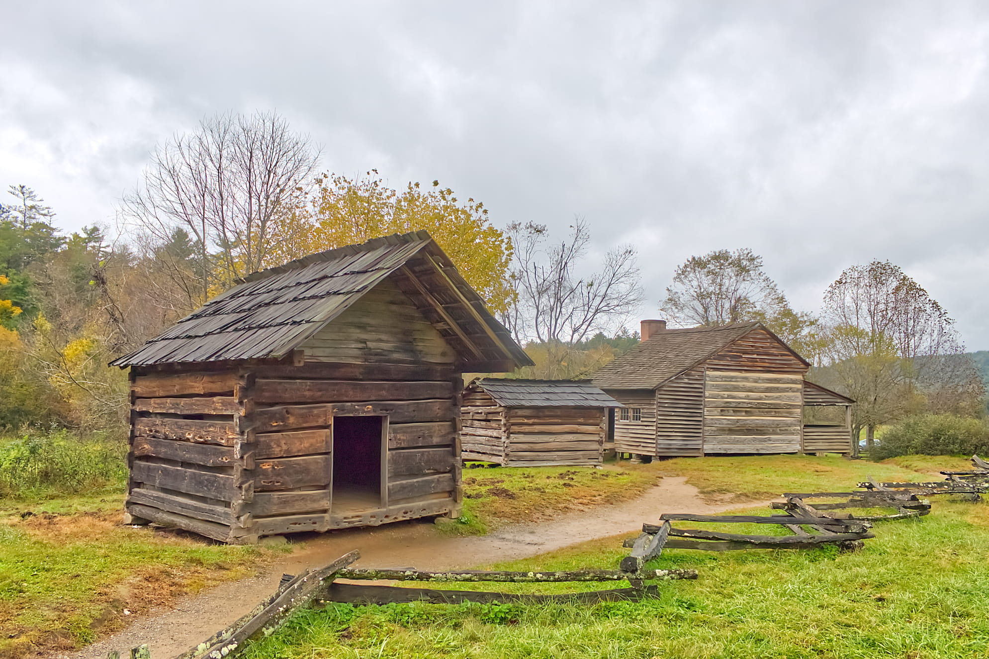 Four historic log cabins with a dirt pathway connecting them under overcast sky. Indicative of civil war era living.
