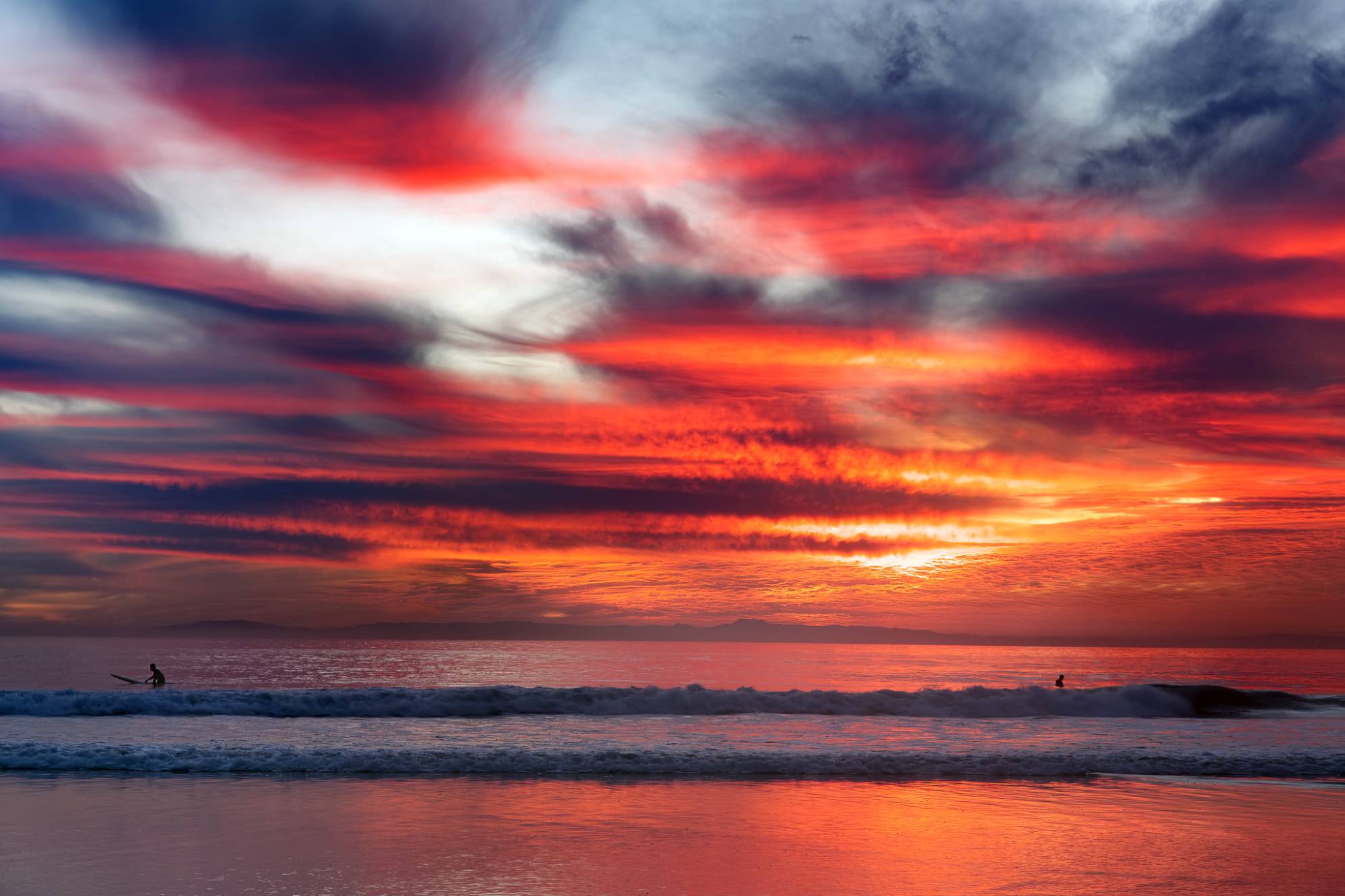 Firey clouds sunset with light blue patch of sky peeking out over reflective Pacific Ocean. Two surfers await waves.