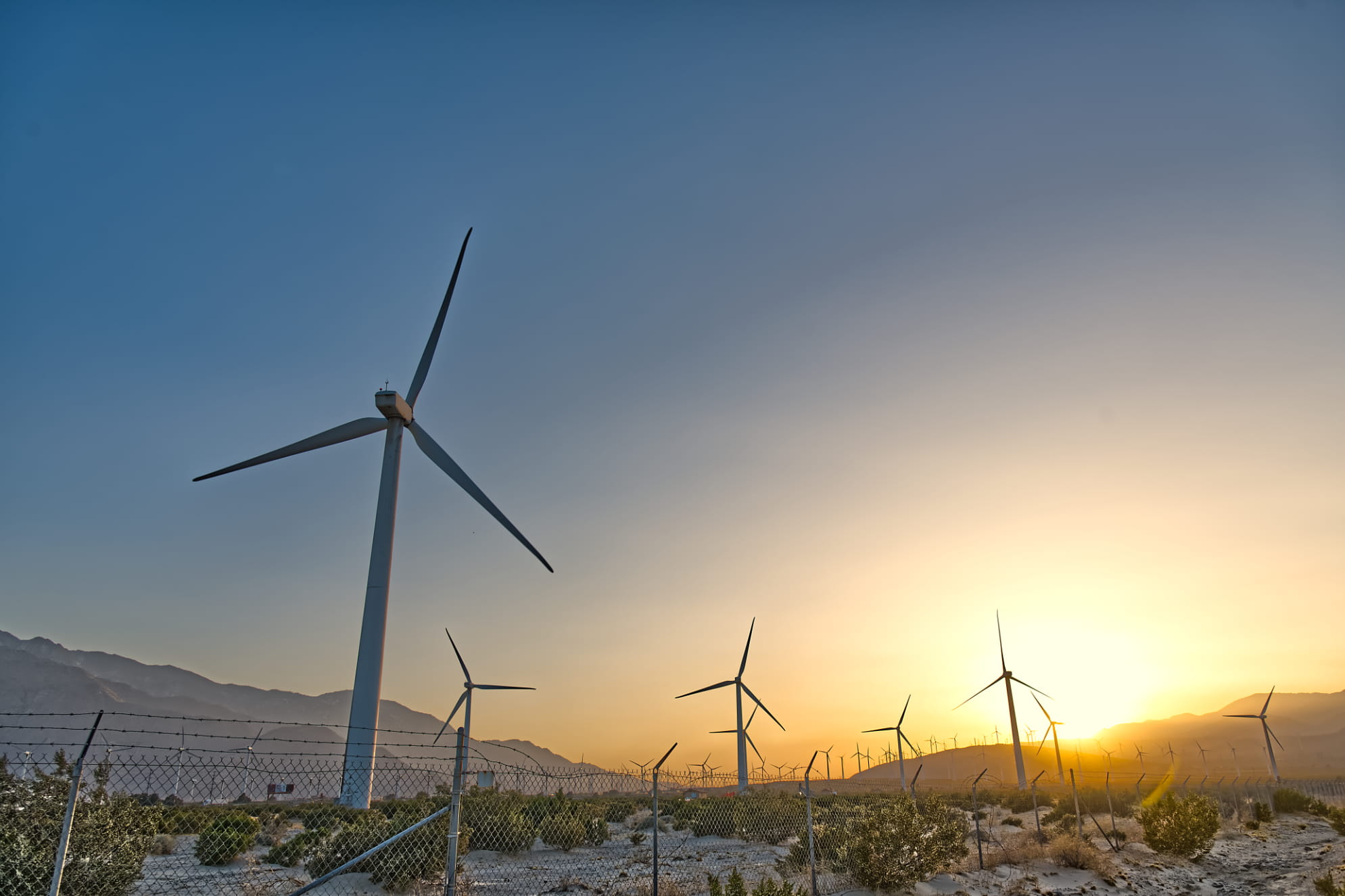 Palm Springs wind turbines captured in stillness afront blue sky fading to golden yellow sunset over mountains.