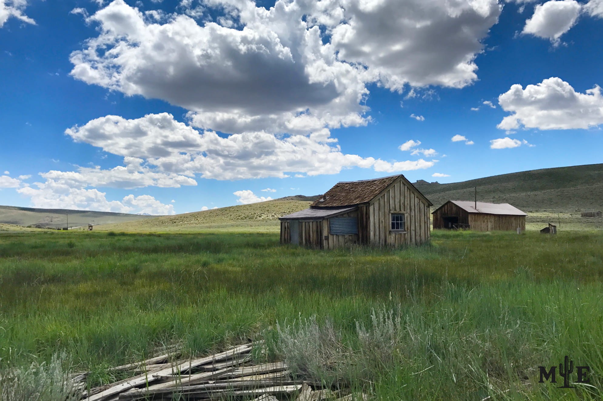 Historic wooden dwelling in Bodie, California, standing on rolling green hills beneath a blue sky scattered with clouds.