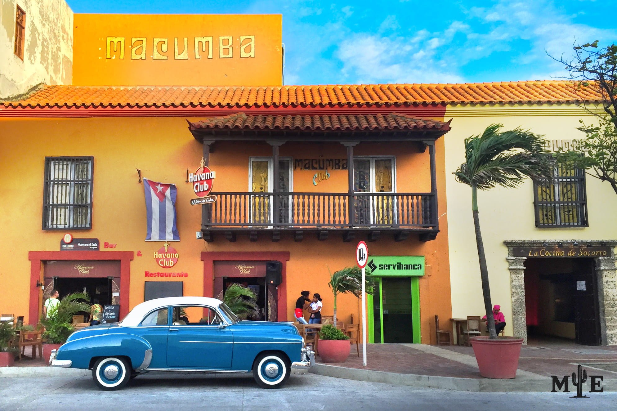 1950s era car in front of a Spanish themed looking building of orange and bronze. Blue sunny day.