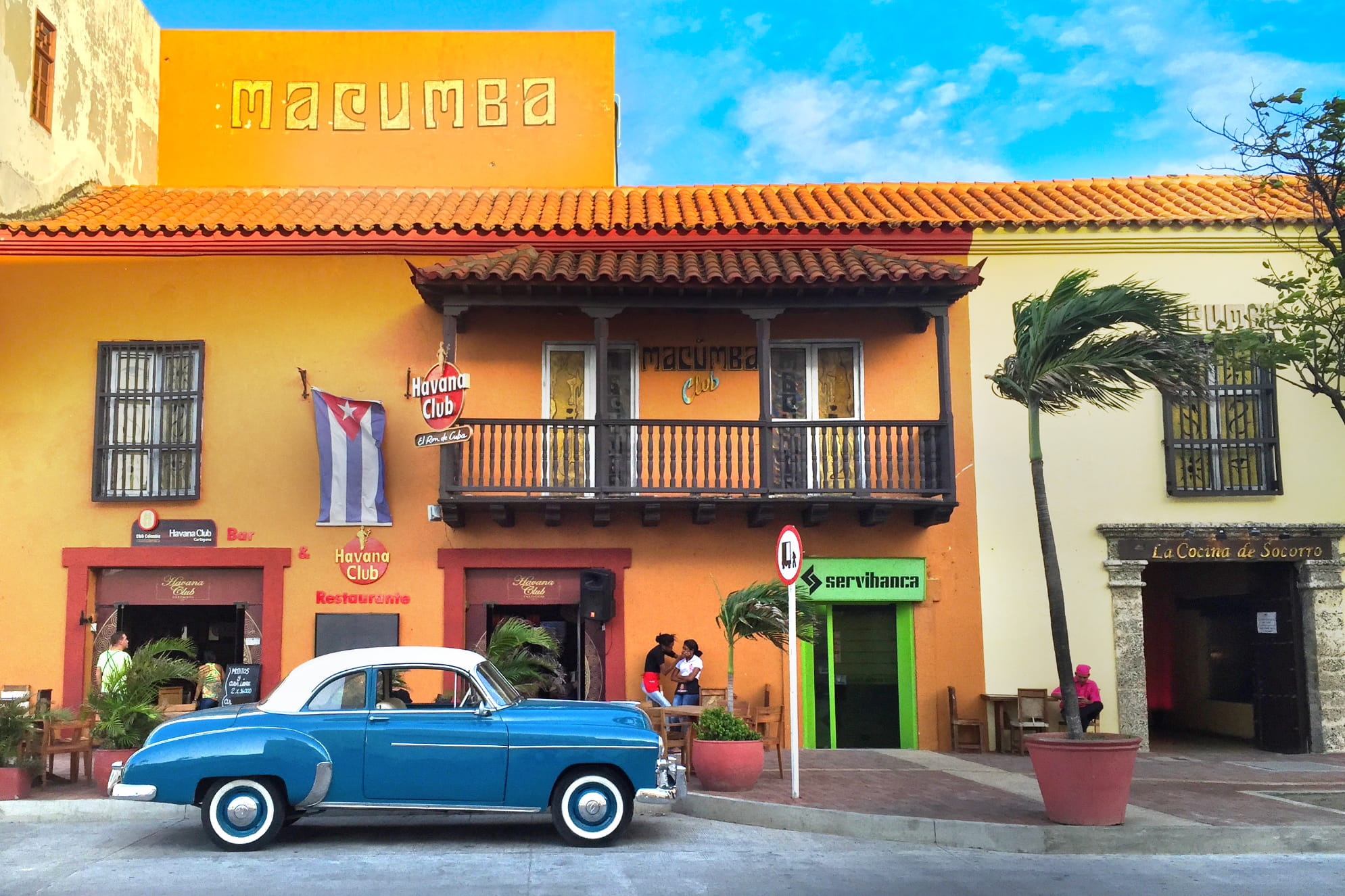 1950s era car in front of a Spanish themed looking building of orange and bronze. Blue sunny day.