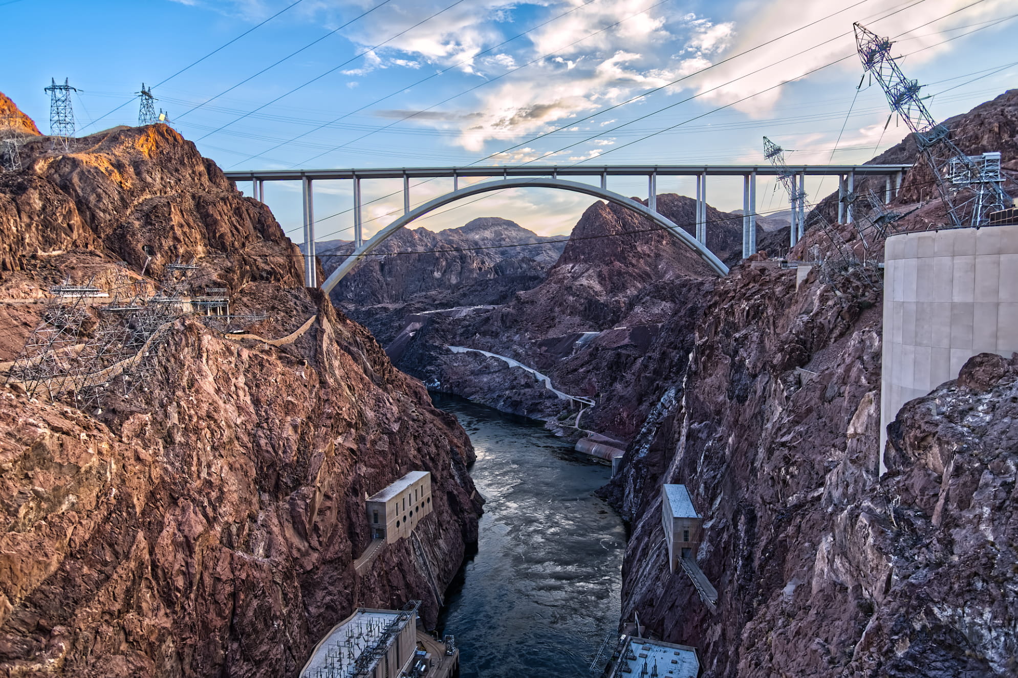 Hoover Dam nestled between canyon walls with the Colorado River below and misc equipment in the mountainside.